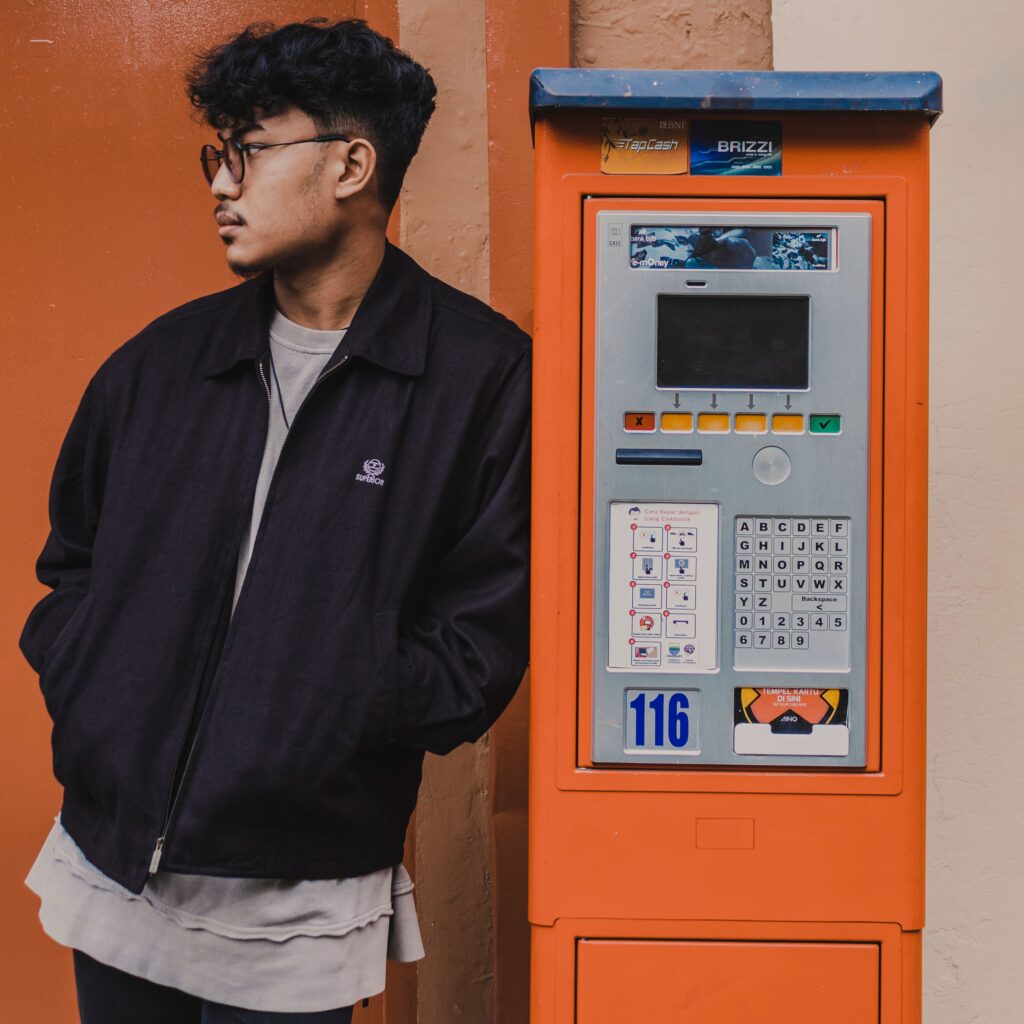Contemplative young ethnic male in casual wear standing with hands in pockets near outdoor payment kiosk machine and looking away in thoughts